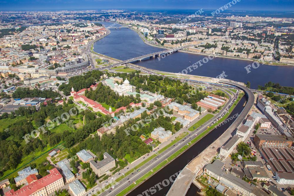 На снимке — панорамный вид Санкт-Петербурга, сделанный с дрона. В центре кадра расположен величественный Смольный собор — один из шедевров архитектуры елизаветинского барокко, окружённый комплексом зданий Смольного монастыря. Виден Большой Смольный мост через реку Неву и современные транспортные развязки. На дальнем плане — центральные районы города, уходящие к горизонту. Снимок отражает гармонию истории и современности в городской структуре Петербурга.